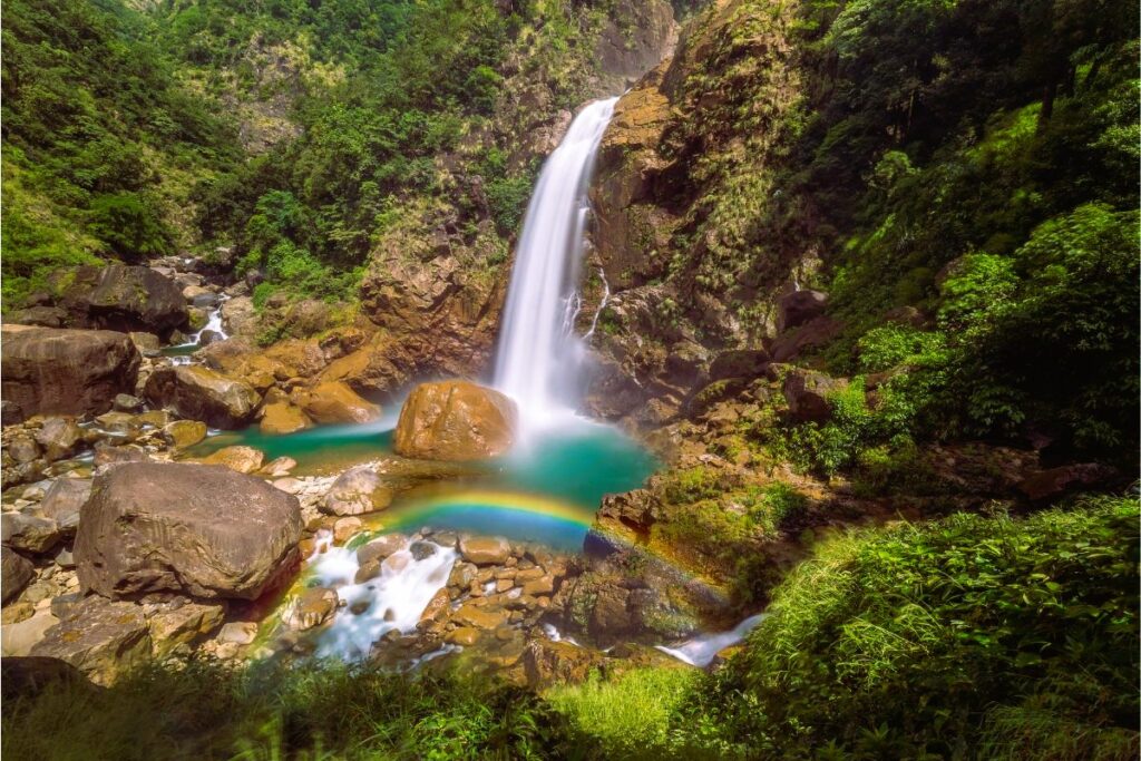 A stunning view of Rainbow Falls in Meghalaya, with the waterfall dropping into a deep blue pool and a natural rainbow forming in the mist